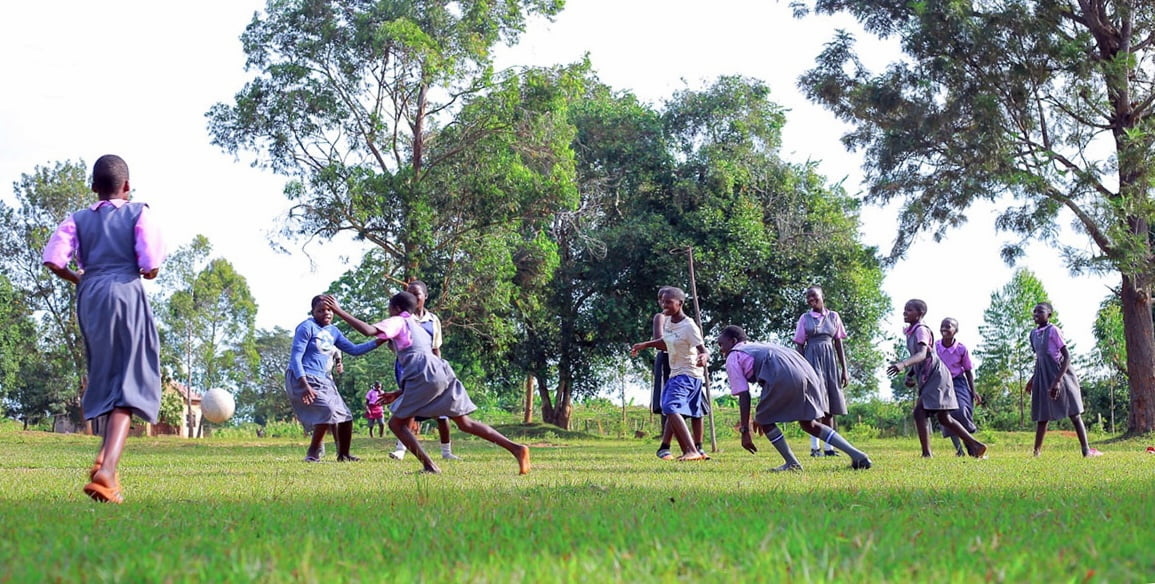 A group of girls in school uniforms playing a ball game on a grassy field, with tall trees behind them.