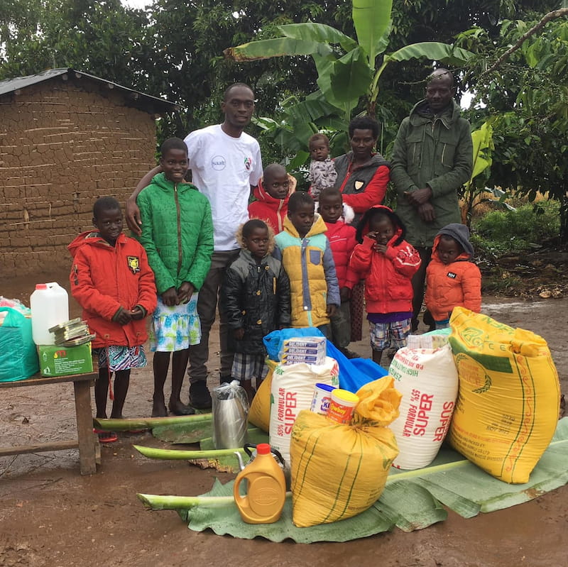 A DROTY staff member stands with a family at their home, surrounded by food, hygiene supplies, and exercise books distributed through the COVID-19 Food Relief Program in 2020.