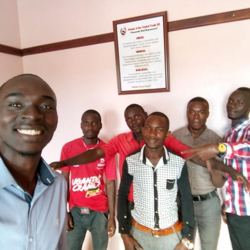 Co-founder Timothy Ssessaazi takes a selfie with team members at a DROTY office, the organization's framed vision statement visible on the wall behind them.