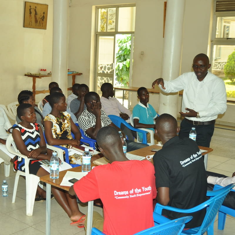 An early DROTY community meeting: young Ugandan adults sit in rows of plastic chairs, listening as a man in a white shirt speaks from the front of the room. Two attendees in the foreground wear DROTY "Community Youth Empowerment" t-shirts.