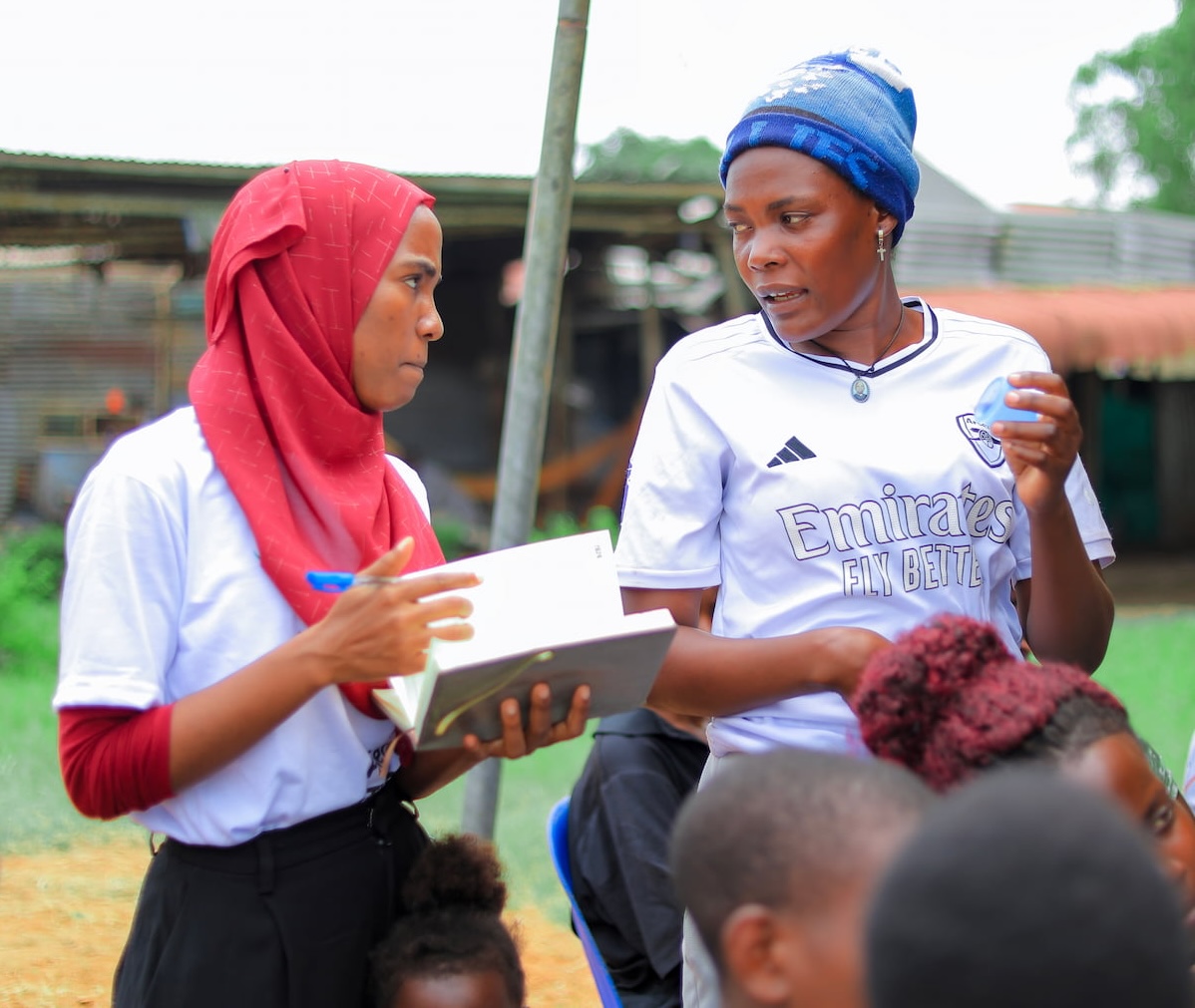 Two young women in casual clothing stand together facing a small audience, one holding a notebook and the other gesturing while speaking.