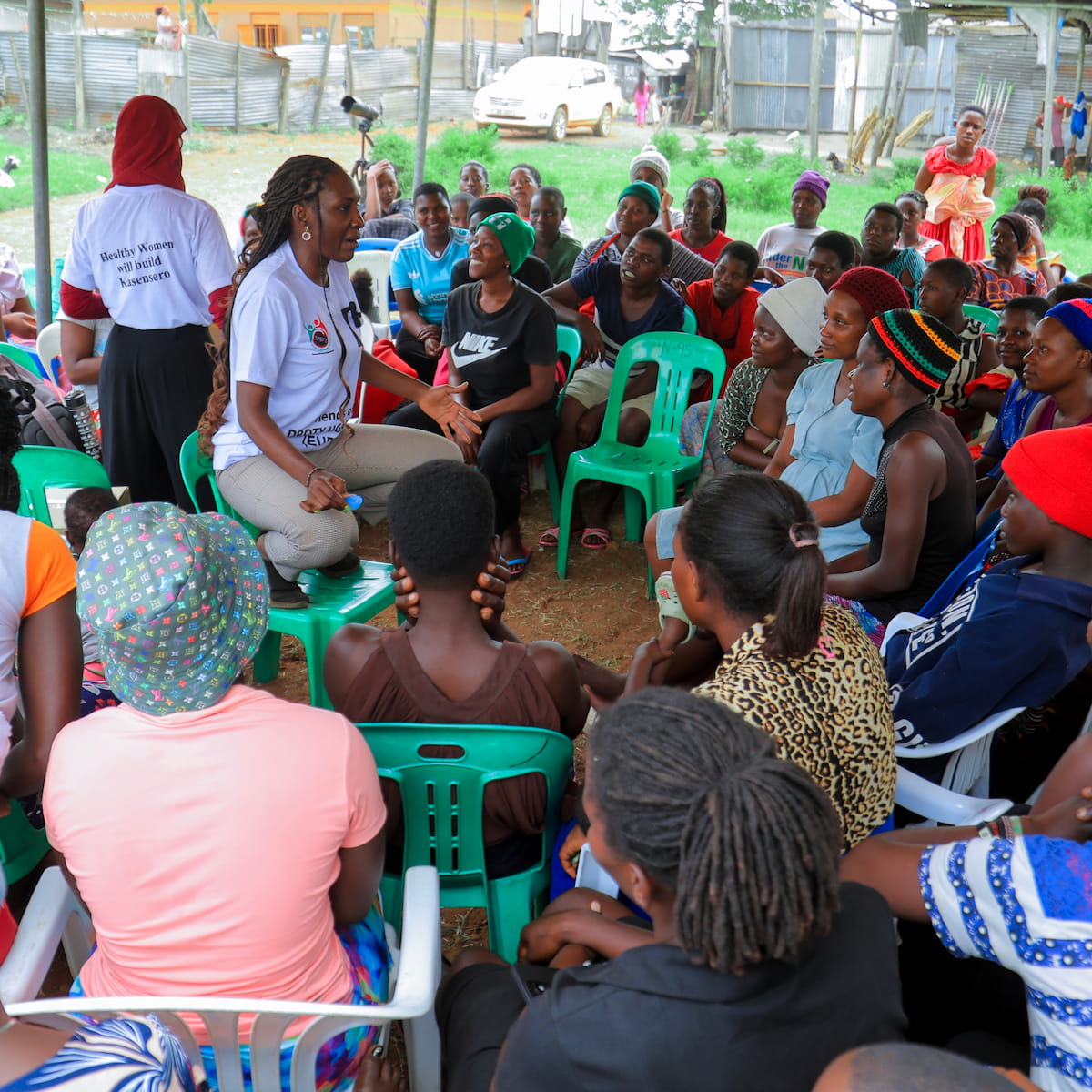 A DROTY staff member sits among a community group of women and girls under a tent, leading a discussion. A second staff member in a "Healthy Women Will Build Kasensero" shirt stands nearby.