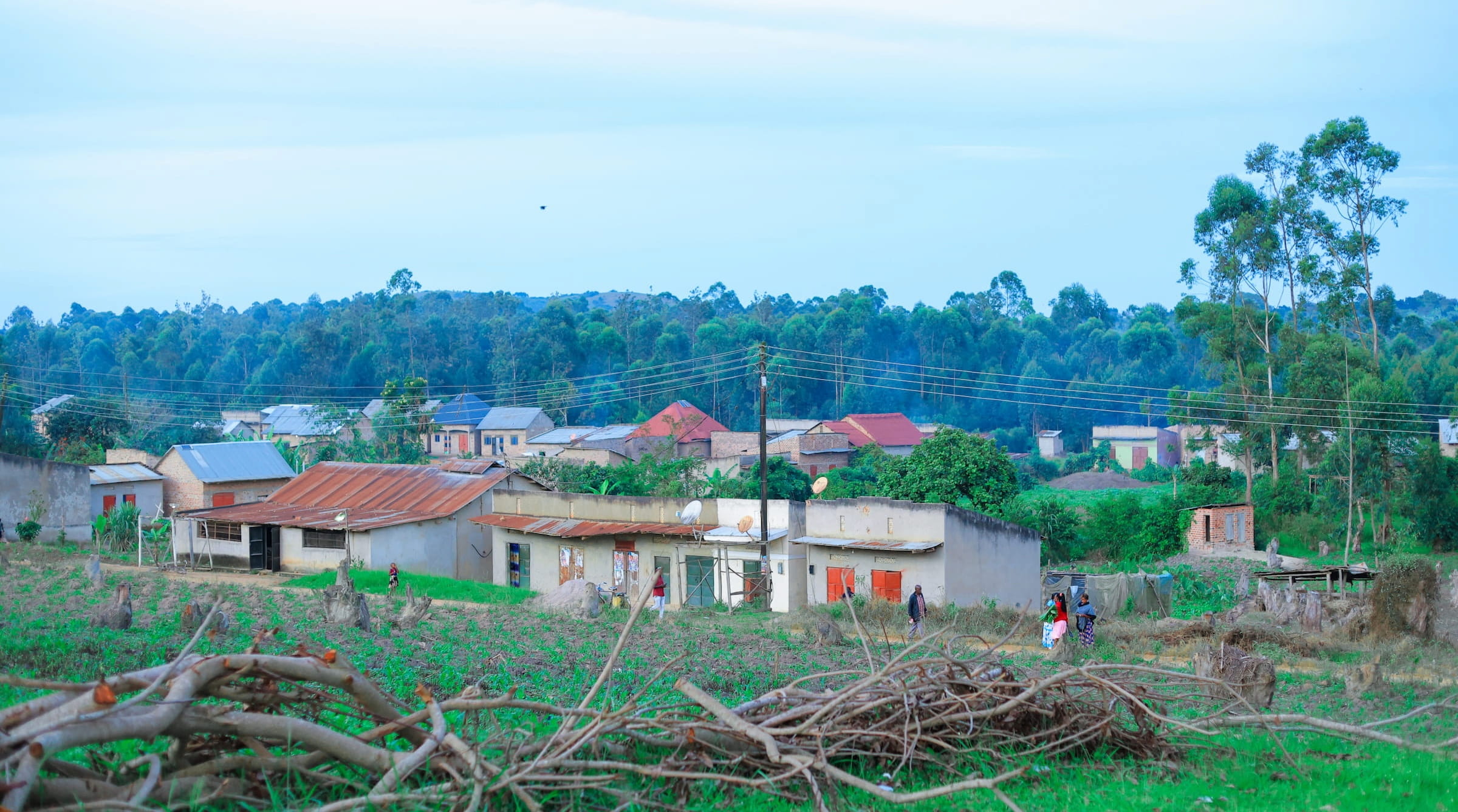 A rural village in Kyotera with rust-roofed homes nestled into a green field, gentle wooded hills behind, with people walking through the foreground at late-afternoon light."