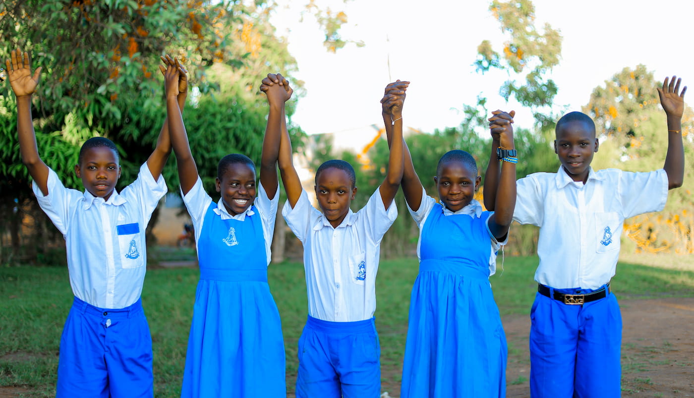 Five children in school uniforms stand in a row with their hands raised and clasped together overhead, smiling at the camera, with green trees behind them.