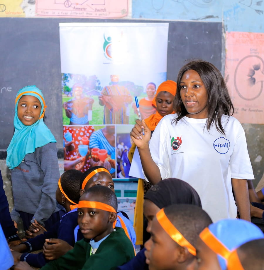 A young Ugandan woman in a DROTY shirt leads a classroom workshop, gesturing with a pen as students in uniform and orange headbands listen.