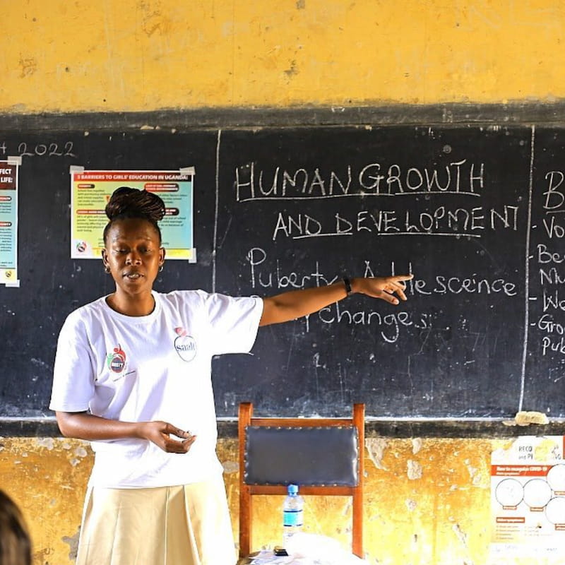 A young Ugandan woman in a DROTY shirt teaches a puberty and human development workshop, pointing to a chalkboard.