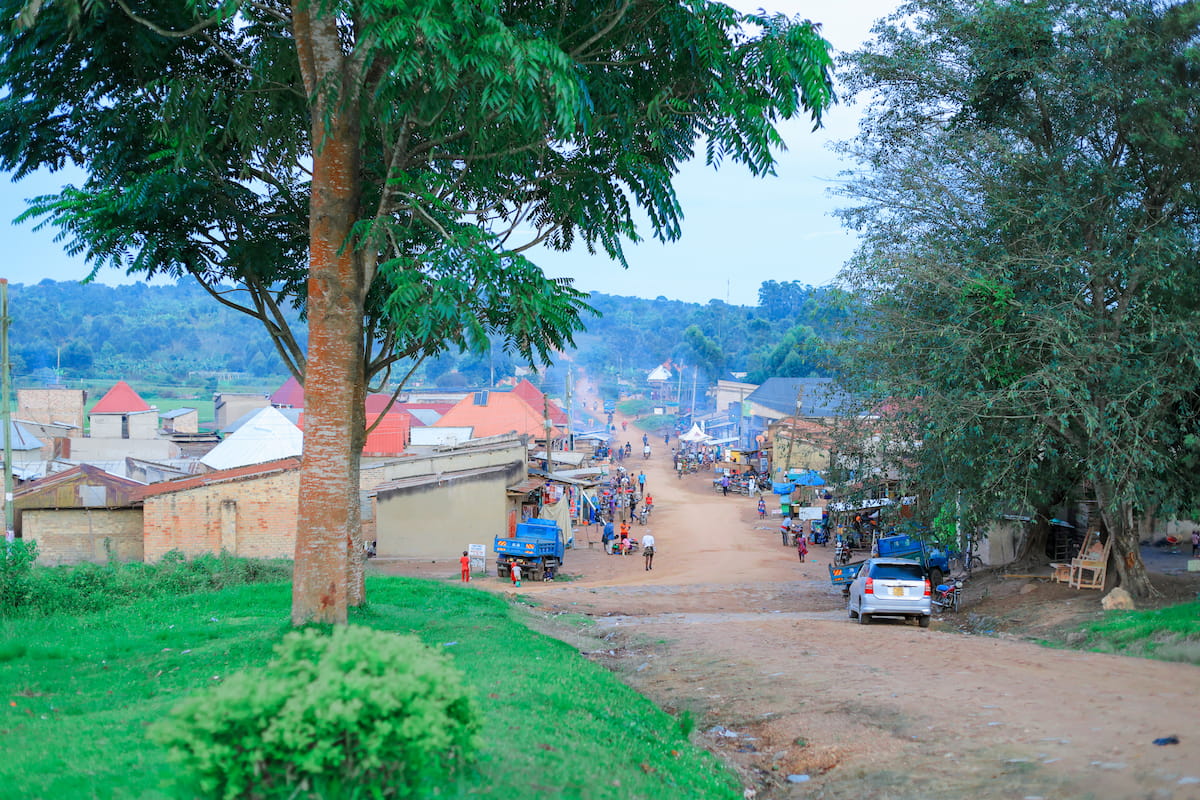 A dirt road winds through a town in rural southern Uganda, lined with small shops and homes with tin roofs, framed by tall trees and rolling hills in the distance.
